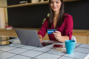 Woman in a red blouse sits at a tiled table using a laptop, holding a credit card—possibly completing 3ds payment processing—with a smartphone, cash, and a blue mug nearby.